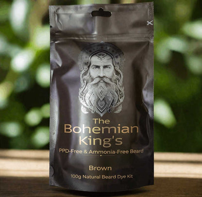 A stand-up pouch package for 'The Bohemian King's' brown beard dye kit, featuring a silver illustration of a bearded king on a dark brown background, set against a blurred green outdoor background.