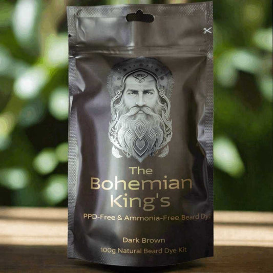 A stand-up pouch package for 'The Bohemian King's' beard dye kit, shown against a blurred green and brown outdoor background. The pouch is dark brown with a metallic silver graphic of a bearded man's face and gold text.