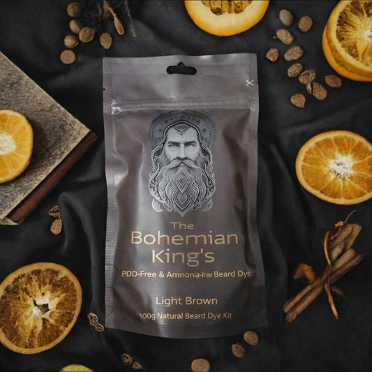 A stand-up pouch of beard dye centered on a dark fabric background, surrounded by dried orange slices, cinnamon sticks, and small brown beads. The pouch reads 'The Bohemian King's' and 'Light Brown,' with a silver illustrated portrait on the front.