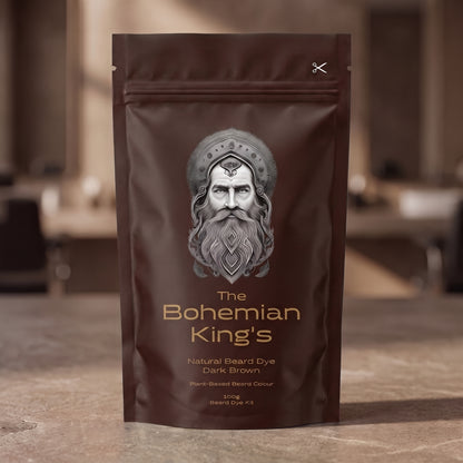 A stand-up pouch of beard dye standing on a stone-like surface with a blurred indoor background. The package is dark brown with a large silver-gray illustrated face and the text 'The Bohemian King's' and 'Natural Beard Dye Dark Brown'. PPD Free Beard Dye