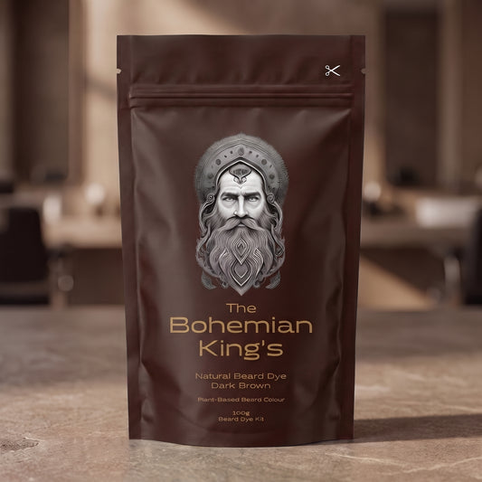 A stand-up pouch of beard dye standing on a stone-like surface with a blurred indoor background. The package is dark brown with a large silver-gray illustrated face and the text 'The Bohemian King's' and 'Natural Beard Dye Dark Brown'. PPD Free Beard Dye