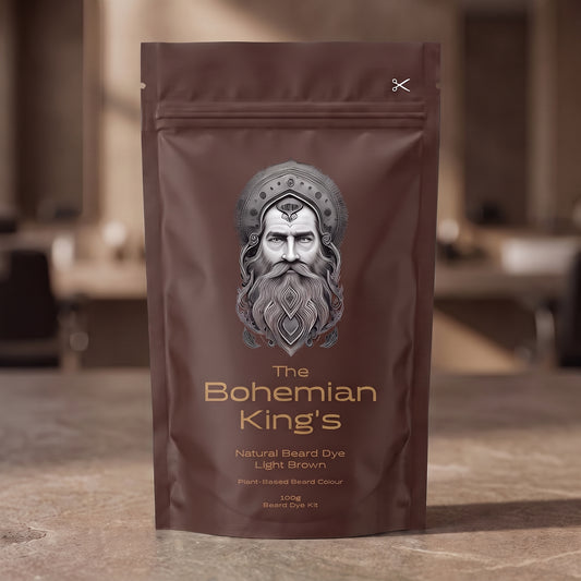 A stand-up pouch of beard dye standing on a stone-like surface with a blurred indoor background. The pouch is dark brown with a large illustrated face and the text 'The Bohemian King's Natural Beard Dye Light Brown' on the front.