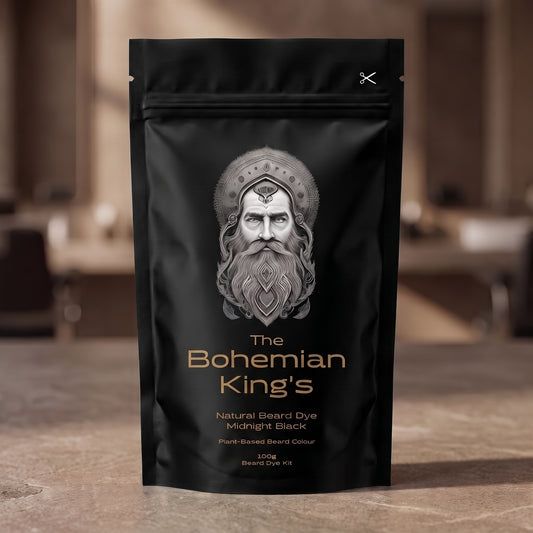 A black stand-up pouch of beard dye standing on a stone surface with a blurred indoor background. The front label shows a silver illustrated beard portrait and the text 'The Bohemian King's Natural Beard Dye Midnight Black' with gold-toned lettering.